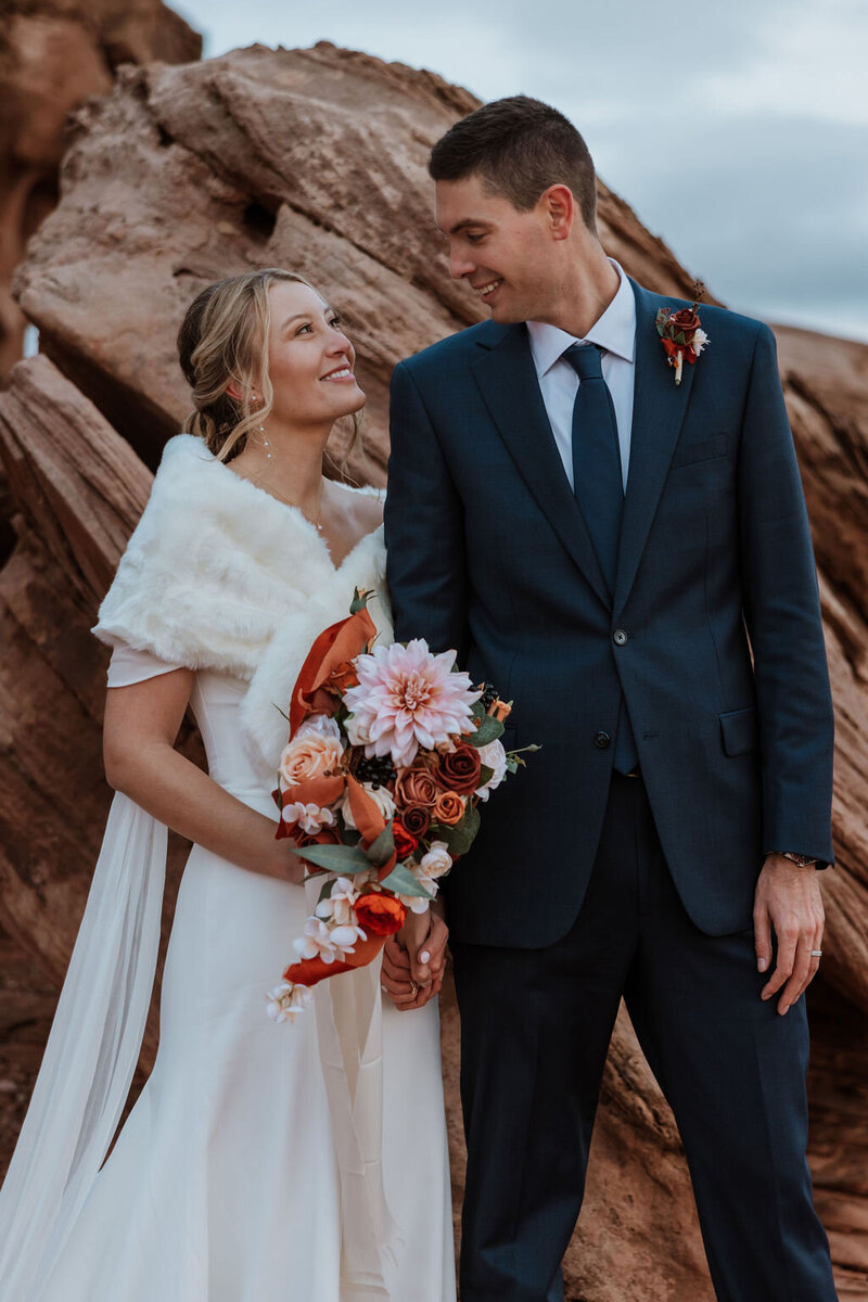 bride and groom standing in the desert in front of large walk wall with rock arch above them