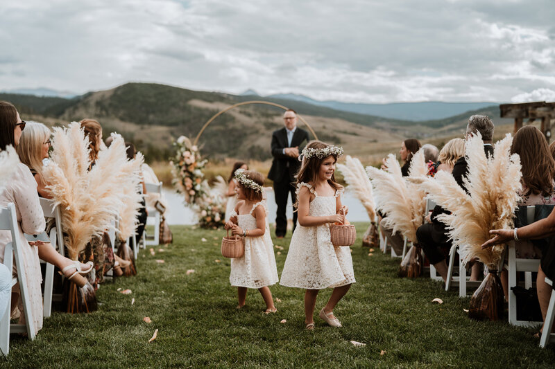 Flower girls walk down a Breckenridge wedding aisle lined with pampas grass at a scenic mountain ceremony.