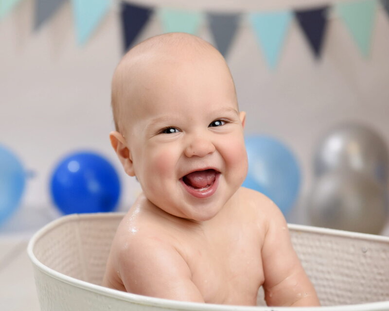 A happy baby with a big smile sitting in a white tub, surrounded by blue and silver balloons and pastel-colored bunting in the background.