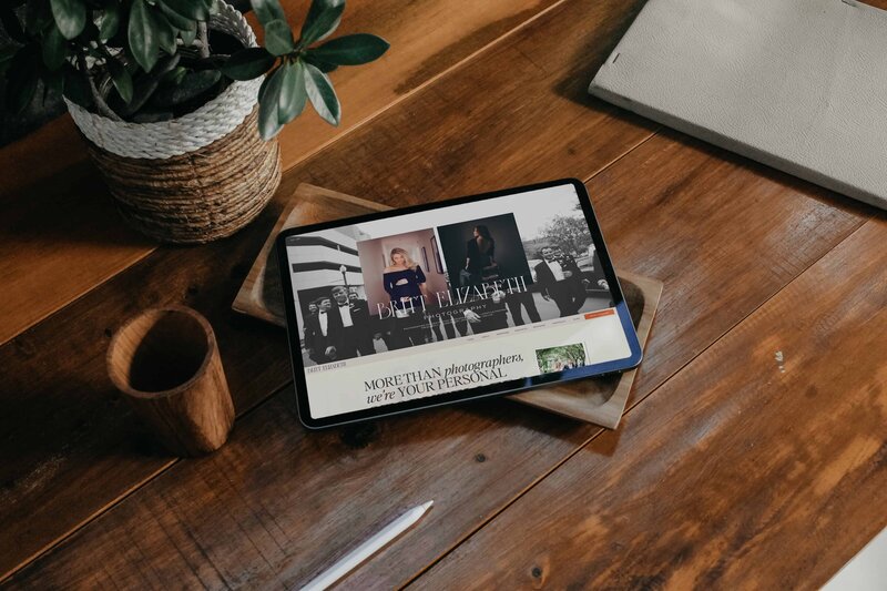 A tablet displaying a website on a wooden table, next to a potted plant, a wooden cup, and a closed notebook with a pen—ideal for those focusing on branding for photographers.