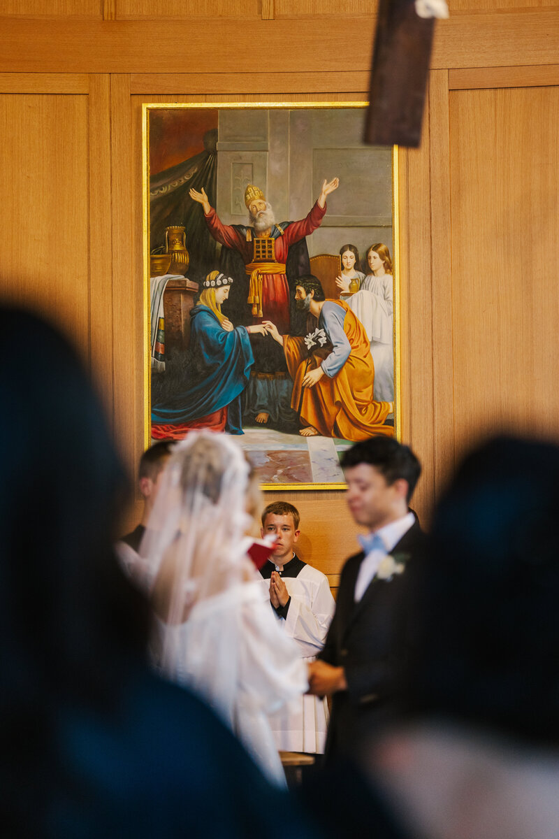 Blurry bride and groom holding hands with priest in focus in the background