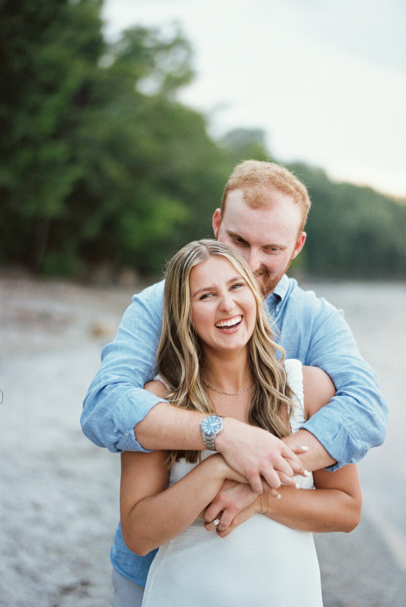 Engaged couple sharing a joyful moment as the man embraces the woman from behind, both smiling on a serene beach with lush greenery in the background.