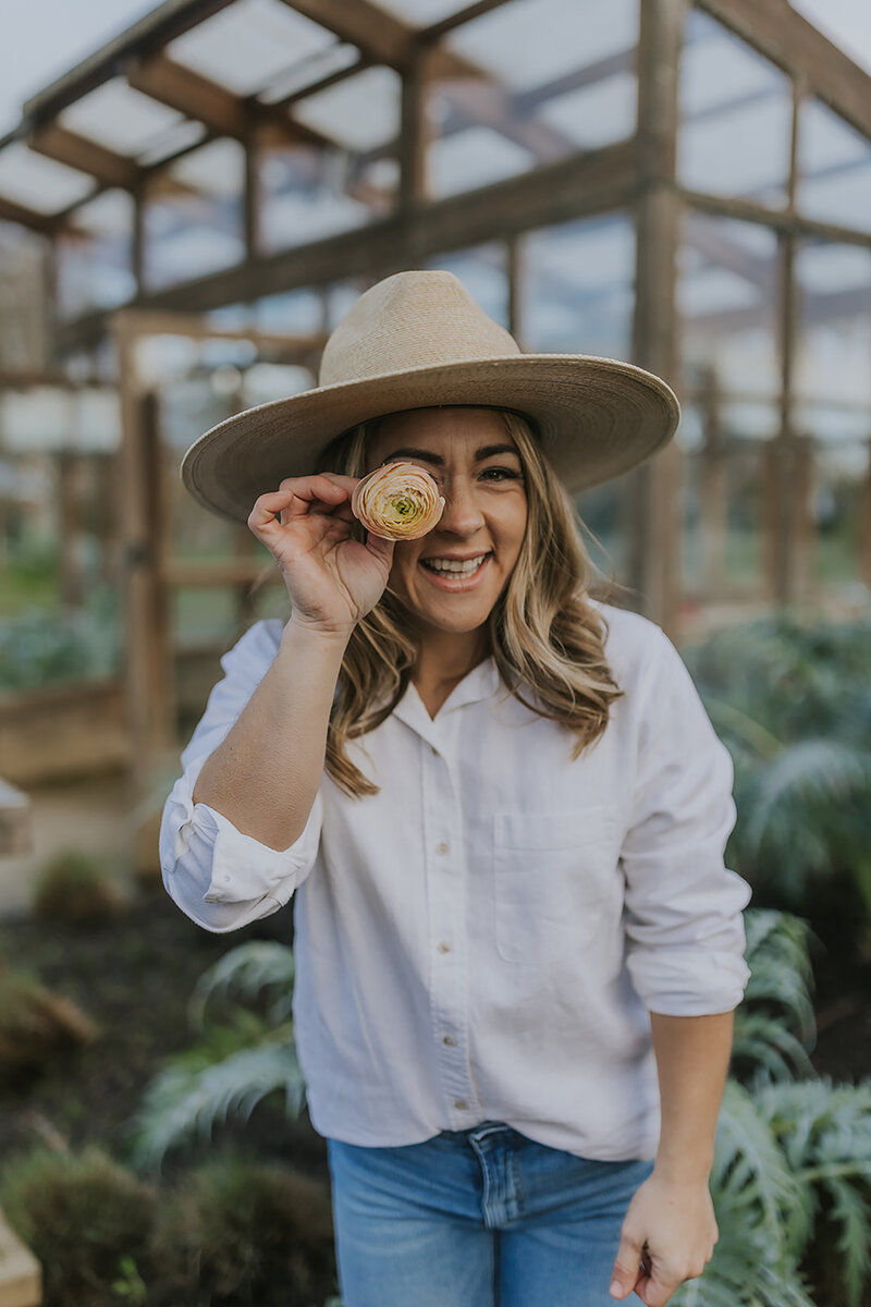 Branding headshot photo for Alexandra wearing cute jeans a white cotton button down and hat while holding a flower up and smiling at the camera