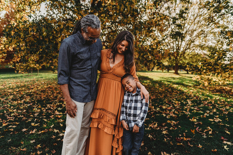 Baltimore photographers captures mother and father embracing one another while the mother holds their child's shoulder and looks to him with the father with autumn trees in the distance for fall family photos
