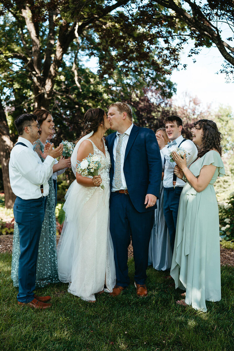 A newlywed couple kissing with their wedding parties clapping around them 
