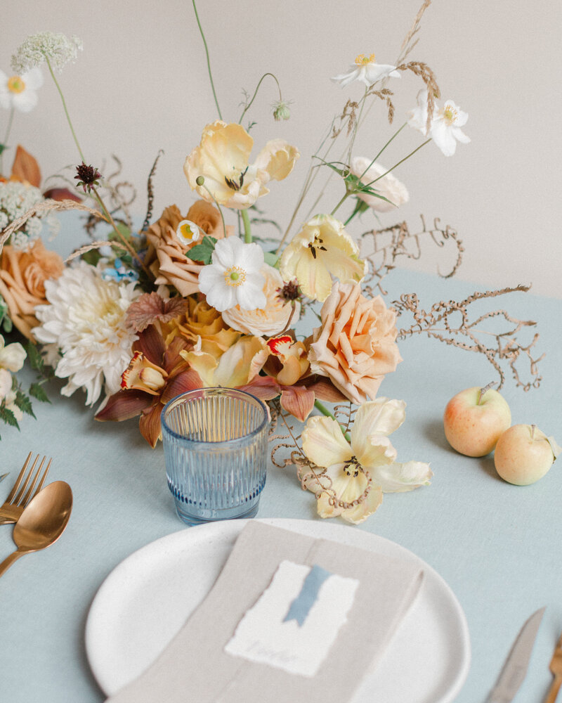 An airy centrepiece of pastel orange flowers sits in front of a dinner table setting. 