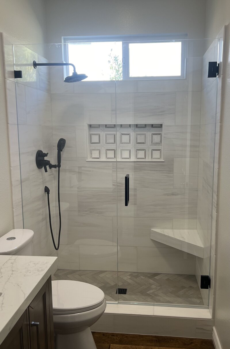 A remodeled shower with a glass enclosure that has black hardware. The tiles are large light grey and white - there is a shampoo box with square tiles.