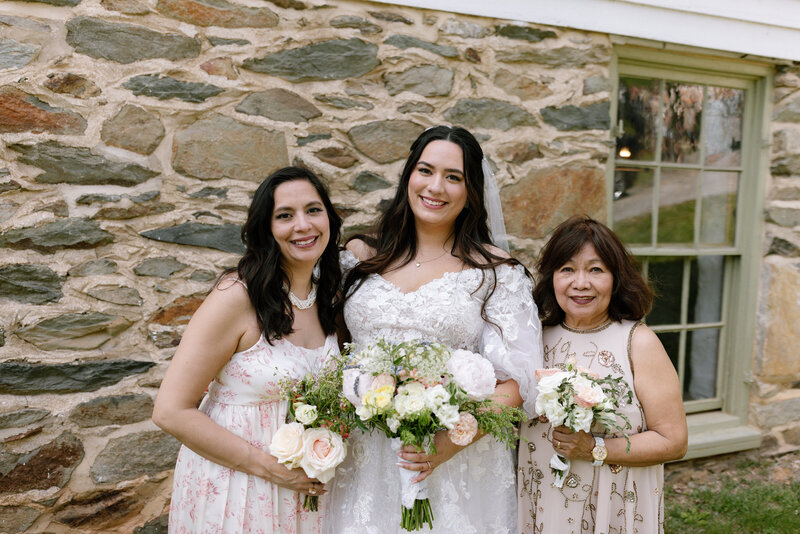a bride with her mother and her grandmother all with dark hair in bridal attire in front of an old stone church  all holding bouquets of roses and peonies