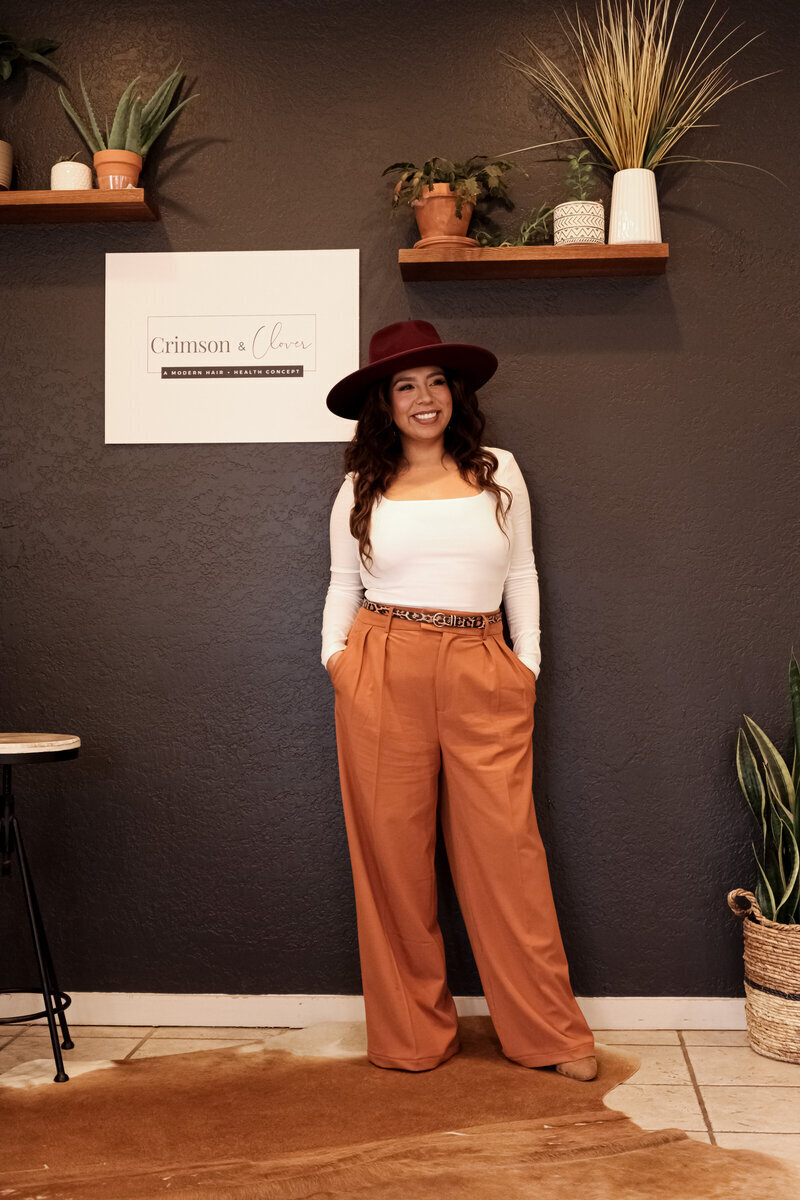 A woman in a wide-brimmed hat, white top, and rust-colored pants stands smiling against a dark wall decorated with plants and shelves. A sign on the wall reads "Crimson &amp; Clover," hinting at an expert touch in websites for salons.