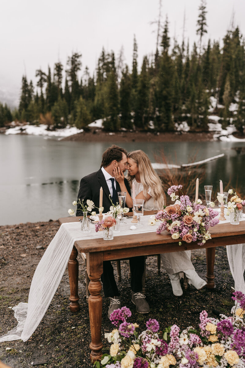 A couple embraces while sitting at their dinner table for two filled with candles and florals.