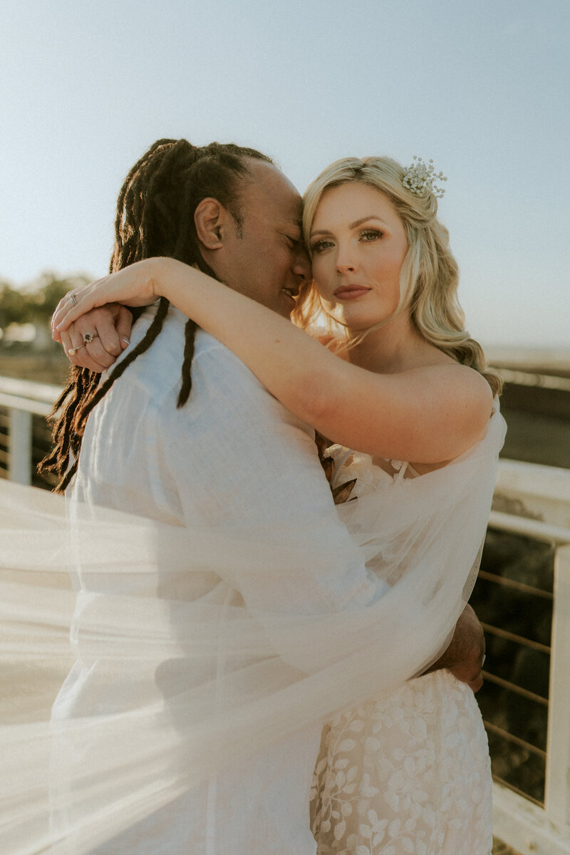 A wedding photo of a Samoan groom and his bride standing on a jetty with her veil sweeping across the shot