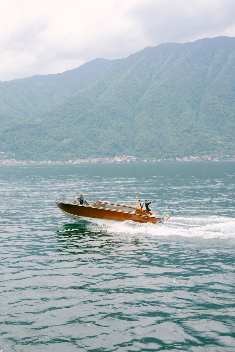 Far away picture of a couple during their wedding wave from a wooden speed boat on lake como in italy