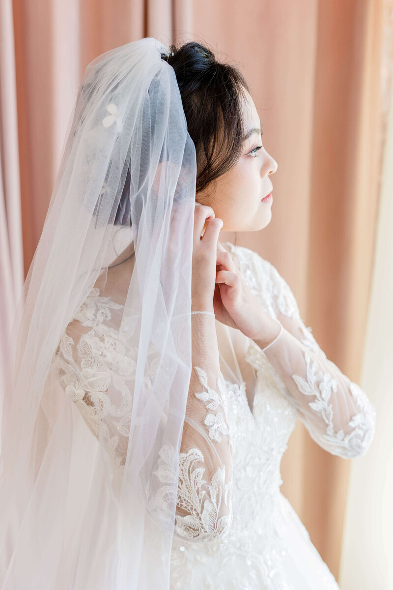 Bride fixing her earring while looking out of a window in a bridal suite on her wedding day.