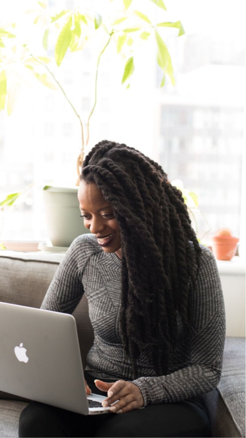 Woman smiling while writing in a notebook outdoors, wearing earbuds and working beside a laptop.