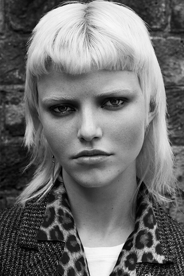 Textured platinum blonde shag haircut with soft layers, styled with a leopard-print collared coat against a brick wall backdrop.