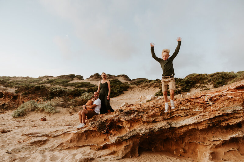 Family standing on cliff top at beach photography session as boy jumps