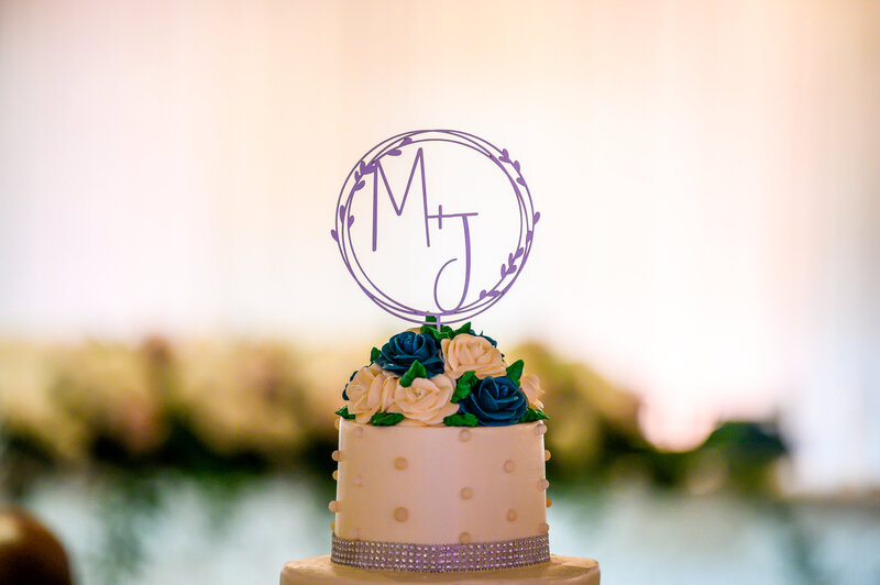 A close-up of a wedding cake topper featuring a custom monogram 'MJ' encircled in a vine design. The topper sits on a light beige cake adorned with a layer of blue and cream roses around the base of the top tier, and a decorative silver beaded border along the bottom of the tier.