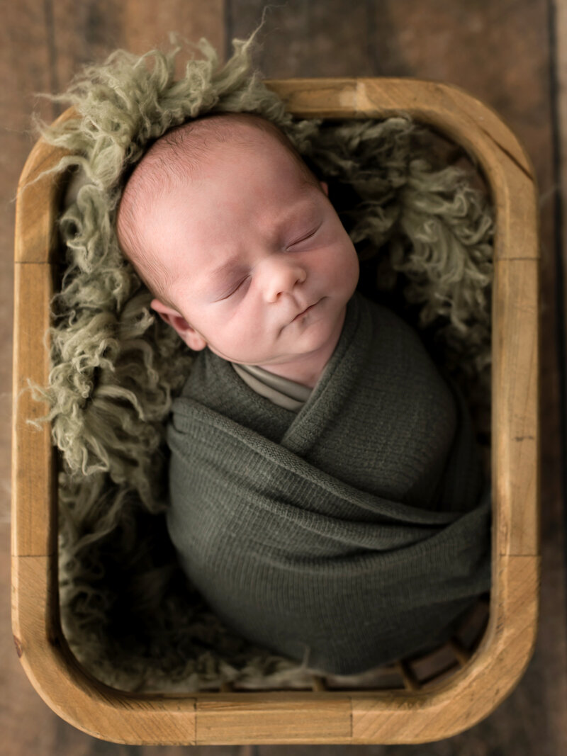 newborn baby boy wrapped in green swaddle posed in basket for newborn studio photoshoot