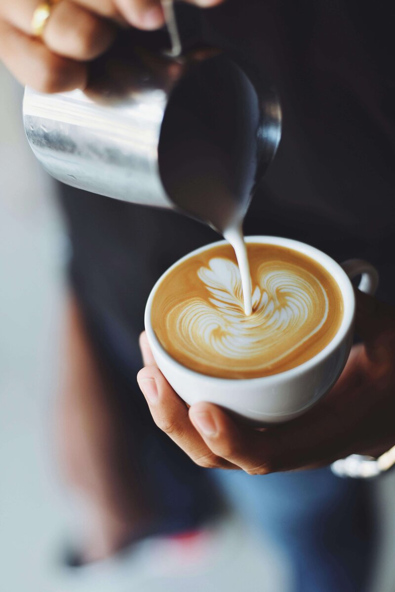 In a cozy coffee shop in Temple, Texas, a skilled barista pours steamed milk into a cup of coffee, weaving an intricate latte art pattern. The soft background blur highlights the artful coffee preparation at its finest.