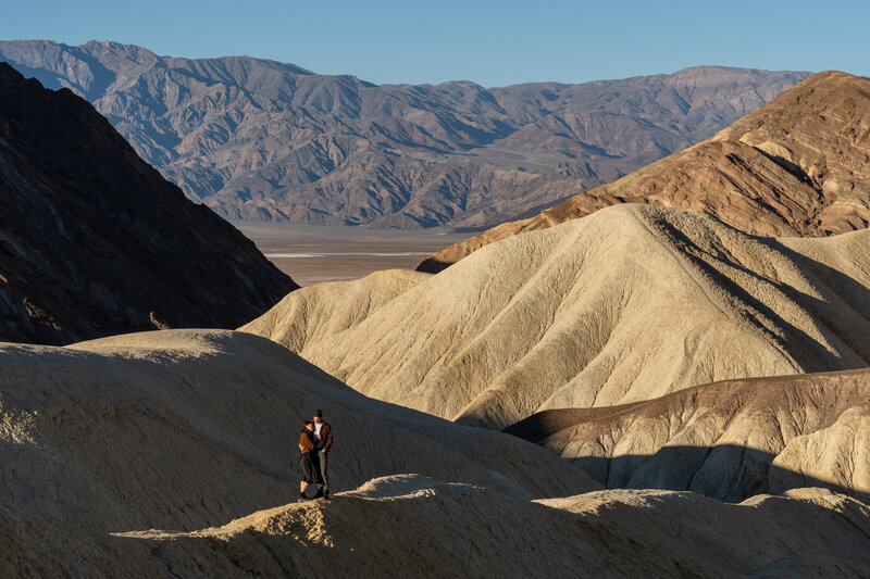 A couple stands on a ridge with a background of mountains and badlands in Death Valley National park