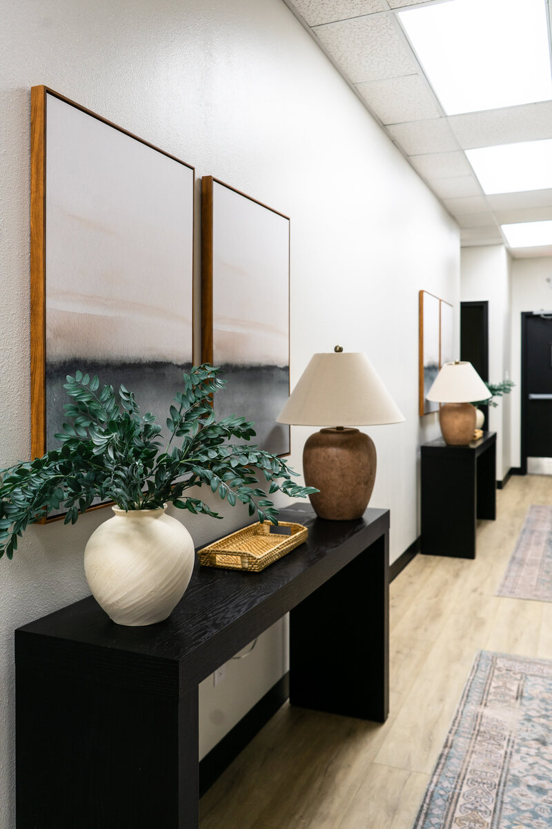 Commercial office space hallway lined with rugs, console tables, lamps, faux plants and artwork.