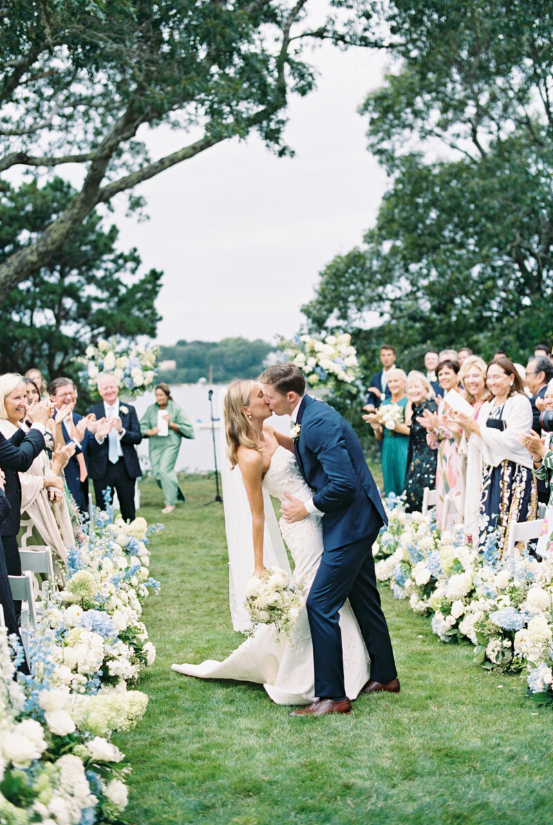 bride and groom stop mid-way down the flower-lined aisle to dip and kiss while guests smile and clap.