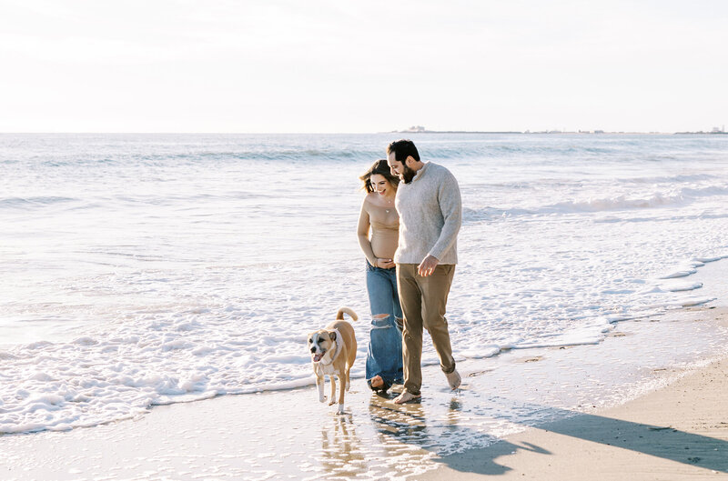 A couple walks with their dog along the surf during their beach maternity session