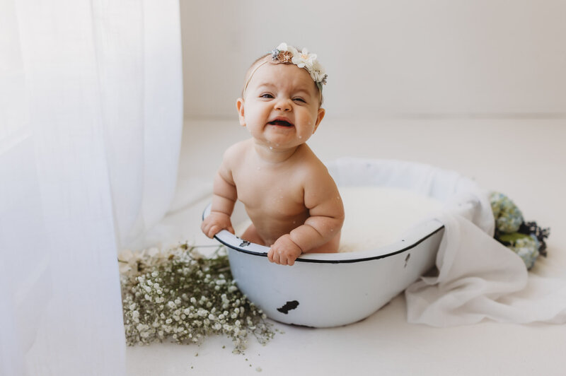 Baby girl in a white bathtub filled with milk, smiling and splashing, surrounded by florals