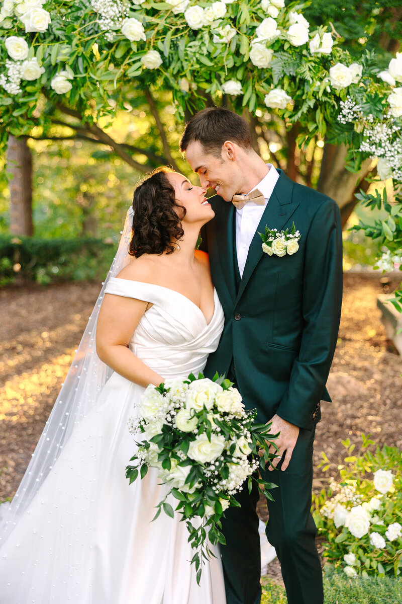 A bride and groom nuzzle noses while smiling as the sun shines behind them through the green trees.