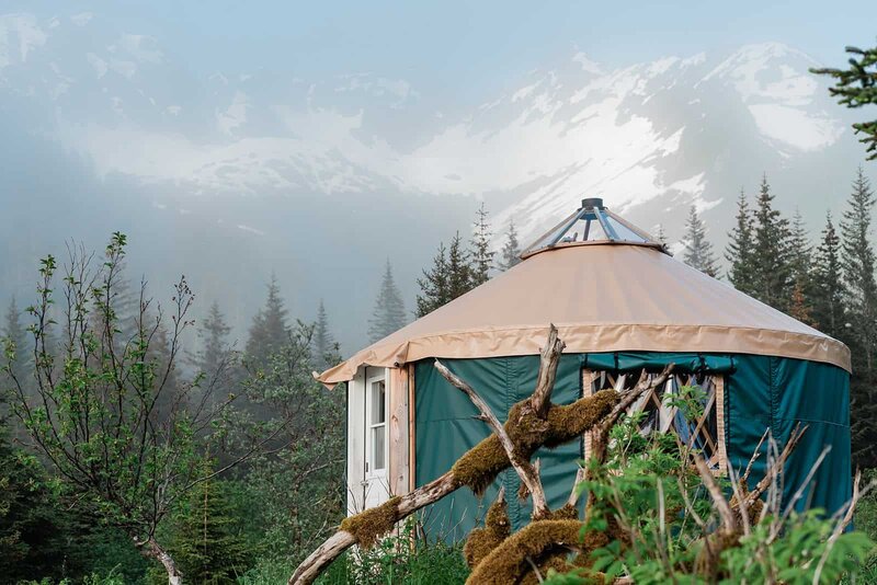 A glamping yurt with light colored roof and forest green walls, is on a platform on the shore of Bear Lake in Alaska where the couple is staying during their two-day luxury helicopter and glamping elopement in Alaska.  There downed tree in the foreground, weather, and covered in lush green moss and green grass all around.  Behind the text is a fog touched view of huge, snow-covered mountains that fill the background.
