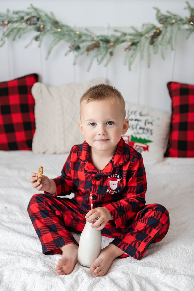 boy on bed in christmas pajamas drinking milk and eating cookies