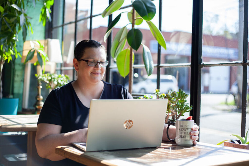 Kristen sitting at her laptop, bathed in sunlight from the large window beside her.