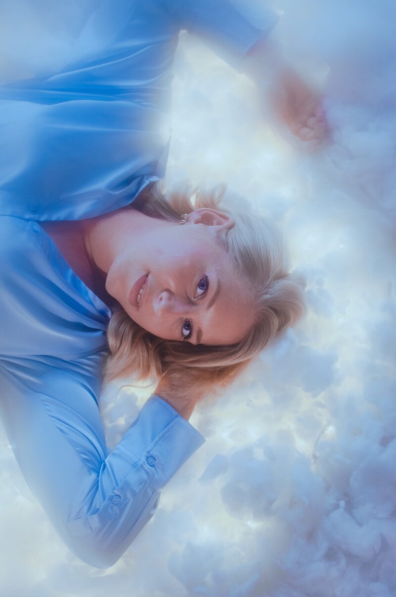 Blonde woman lays against lit up cloud background wearing a blue satin shirt and smiles softly at the camera with her hand resting behind her head.