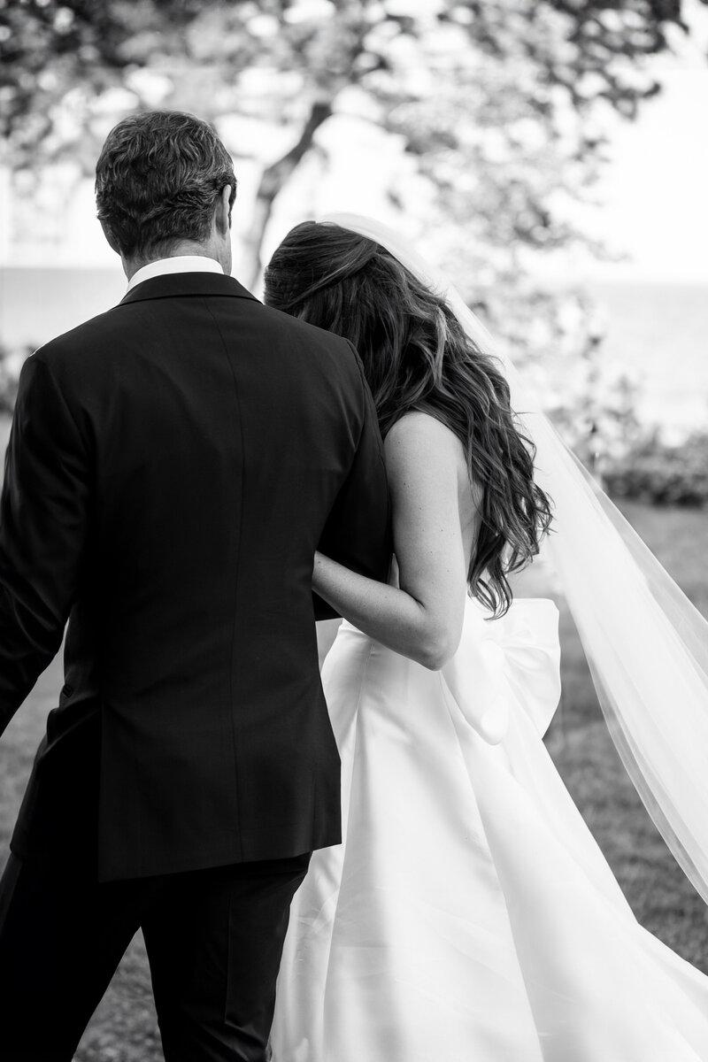 A black and white photo of a bride and groom walking away from the camera. The bride wears a long dress with a flowing veil, holding the grooms arm. They appear in a garden setting with blurred foliage in the background.