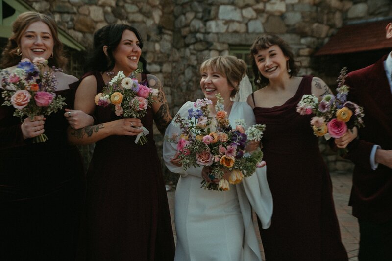 Bride and bridesmaids laughing and posing with garden-style bouquets in front of a stone wall, holding colorful arrangements in mauve, peach, and blush.