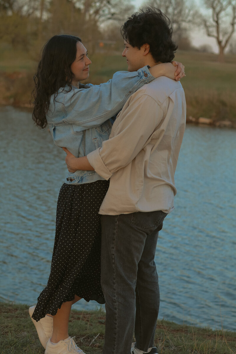 A couple is holding each other and looking at each other fondly. There is water and trees in the background