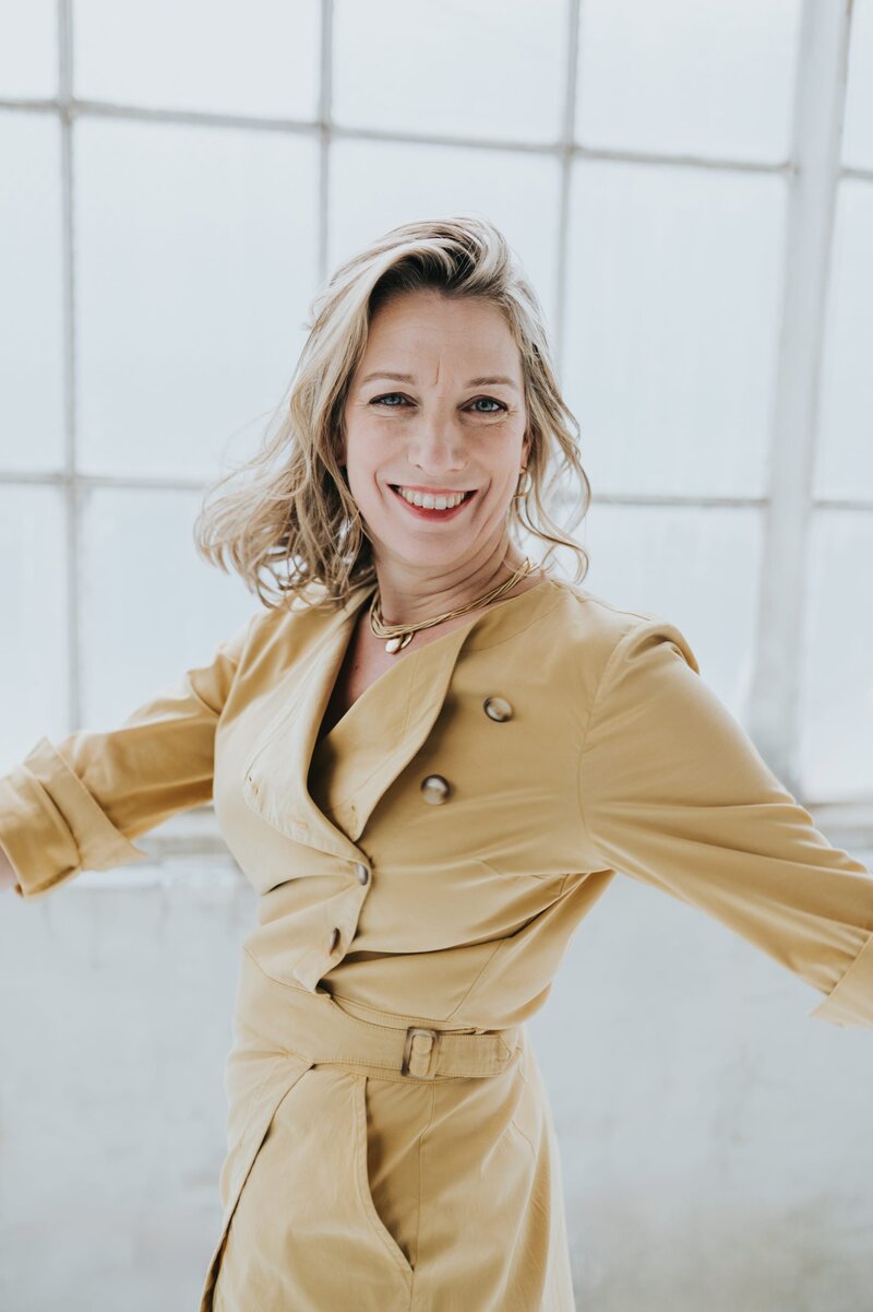 Blonde woman wearing tan dress throws her hands besides her and dances while smiling at the camera.