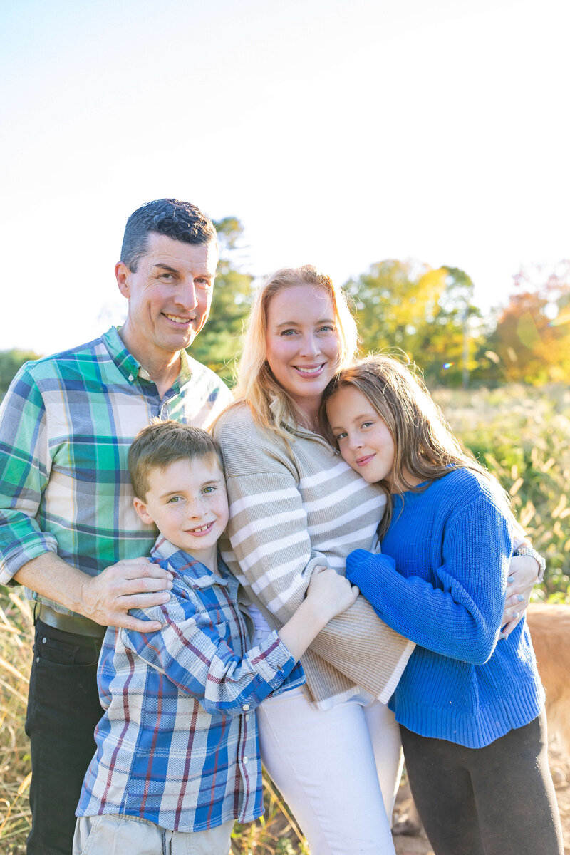 family of 4 photo wearing blue shirts smiling for the camera and hugging mom