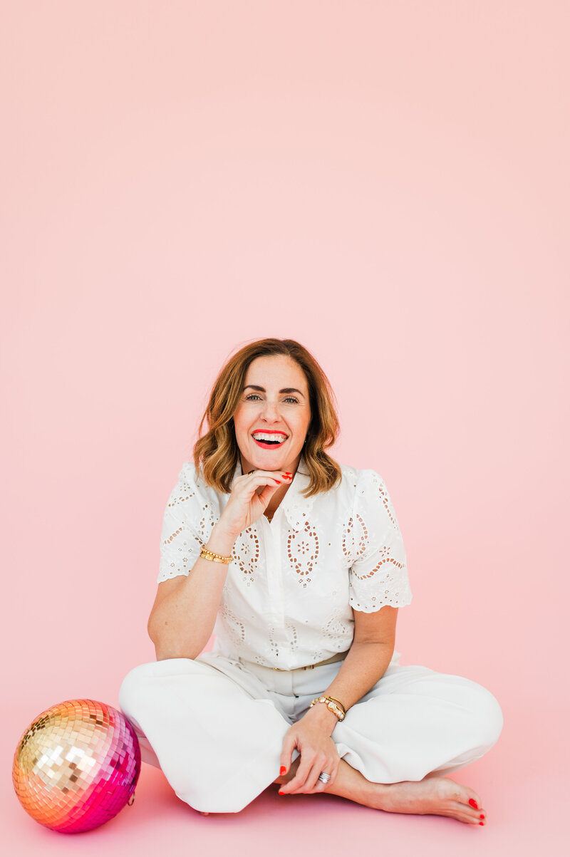 female photographer wearing pink pants and white top sitting next to a disco ball
