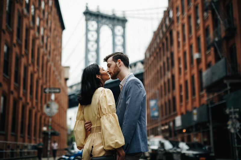A man and woman stand facing each other and touching foreheads on a cobblestone street, with the Manhattan Bridge in the background between two rows of red-brick buildings.