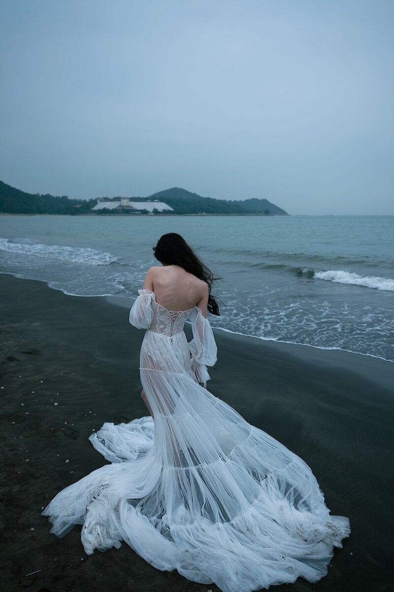 A woman wearing a delicate sheer couture dress shot on the beach in dark moody tones.