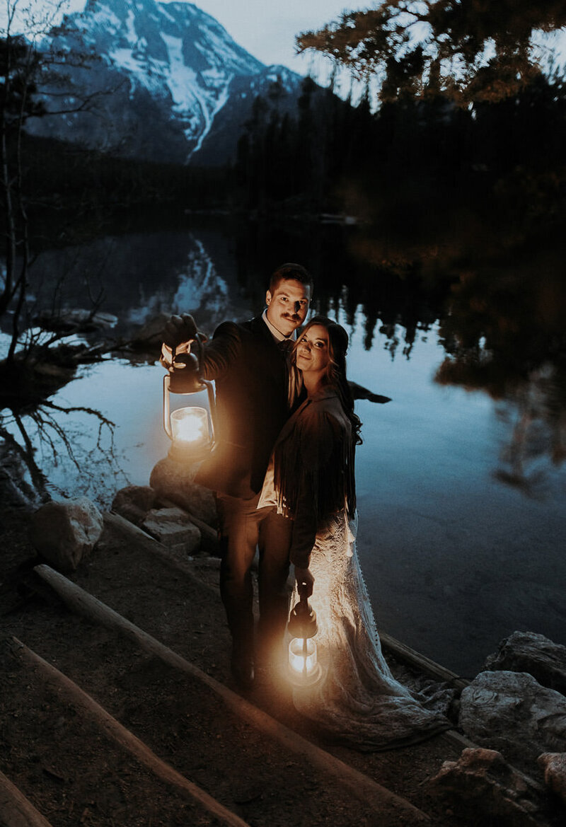 An elopement couple at Leigh Lake in Grand Tetons National Park near Jackson Hole, holding lanterns during blue hour and looking at the camera.