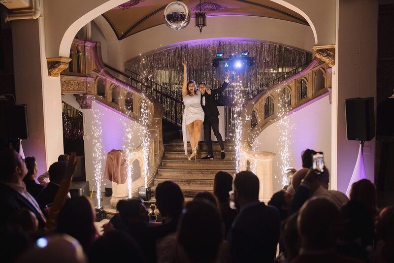 A couple stands on a staircase under a disco ball with indoor fireworks going off at their new years eve wedding reception
