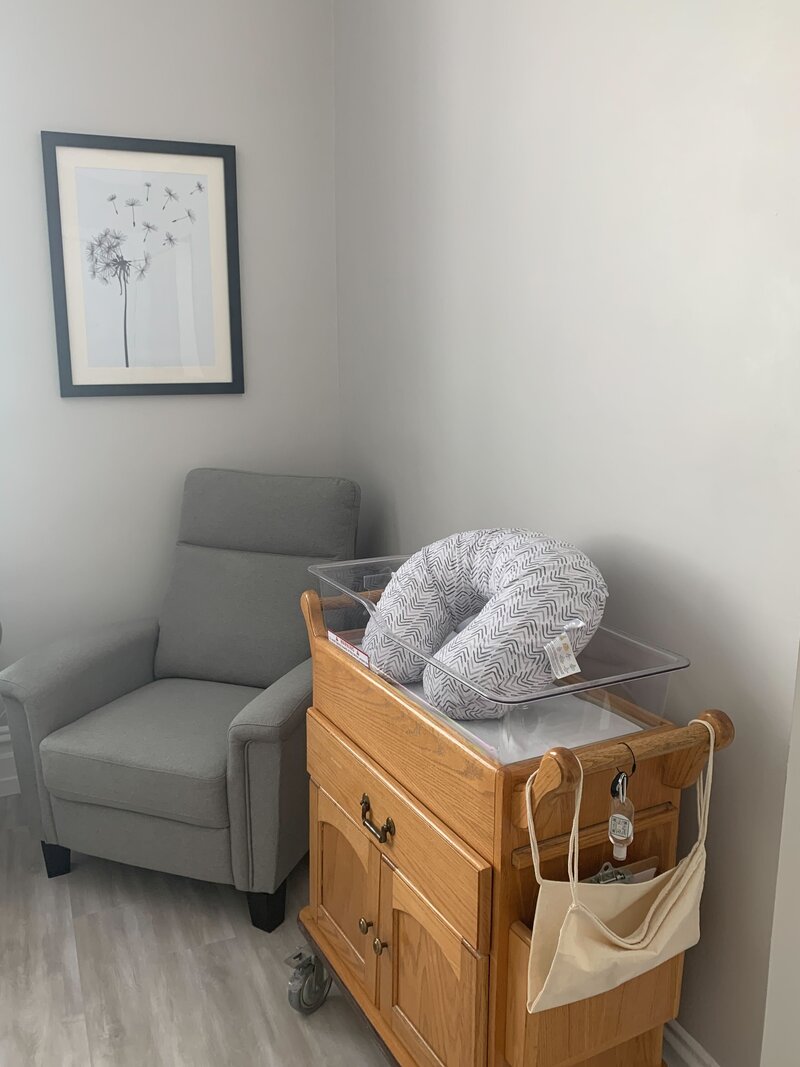 A corner of a room with a gray armchair and a hospital-grade bassinet with a boppy in it.