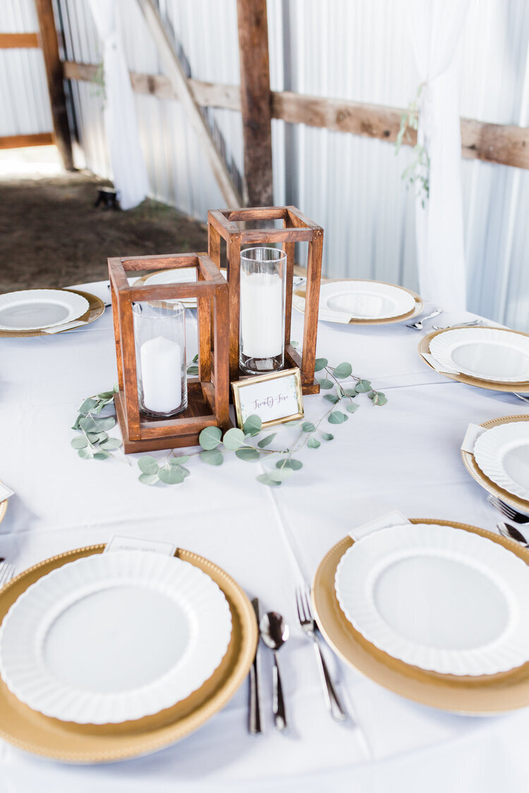 rustic wedding tablescape with wooden lanterns and greenery and white table cloth with gold chargers and white plates coordinated by cait and co events