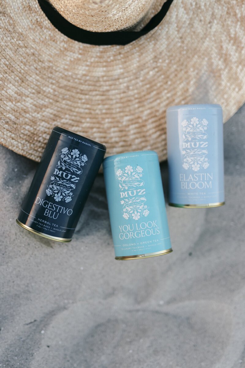 MUZ Wellness Collection consists of three blends: Digestivo Blu, Elastin Bloom, You Look Gorgeous. All blends are created with a focus on wellness and health. Tea cans are seen on a beach along with a sun hat behind them. 