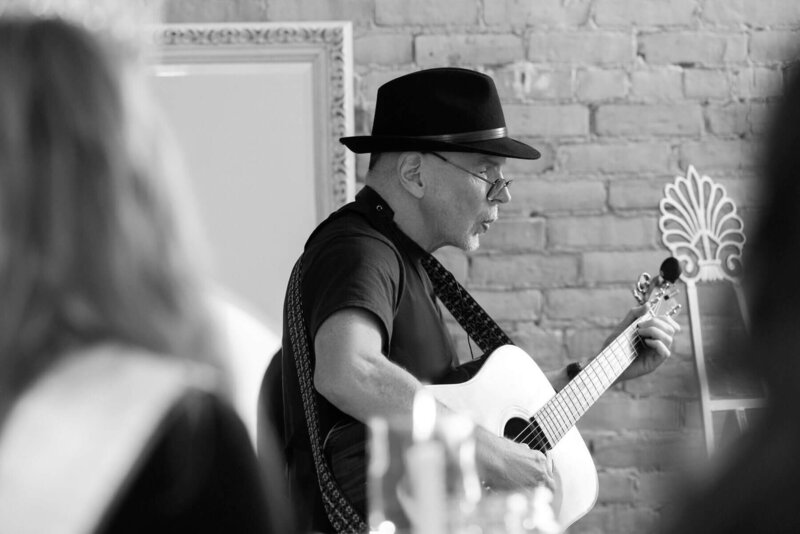 A musician wearing a fedora plays the guitar in a stylish event space with vintage decor.