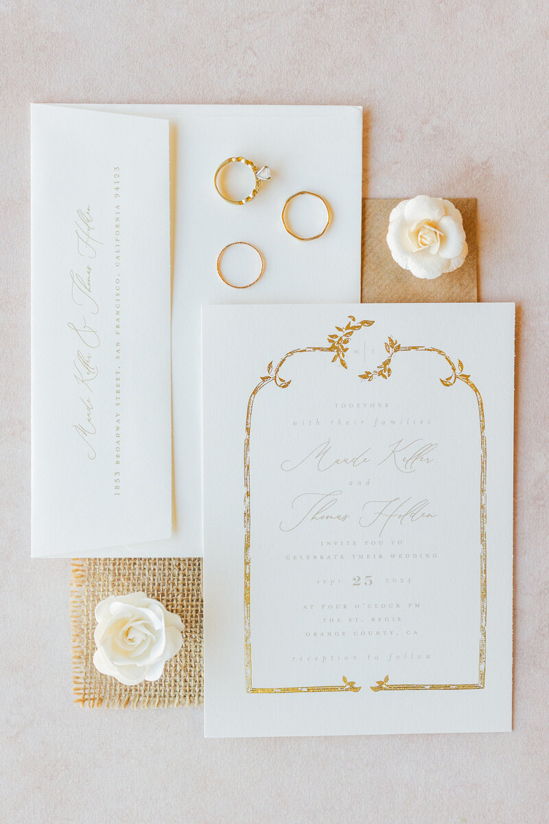 Wedding details with gold trim and rings sit on a white table with white flowers