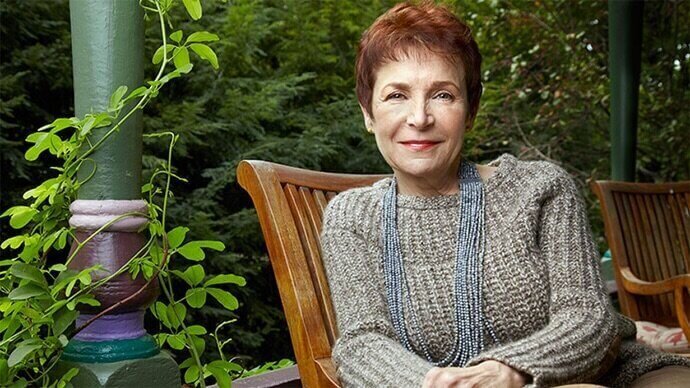 Caroline Myss sits on a wooden chair on a porch, wearing a cozy sweater and long beaded necklace, surrounded by greenery.