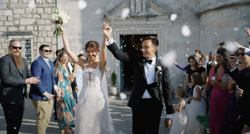 Bride and groom walk out of a stone church as flower petals fly in the air, surrounded by smiling guests during a sunny wedding celebration in Montenegro — moment from an elegant destination wedding film